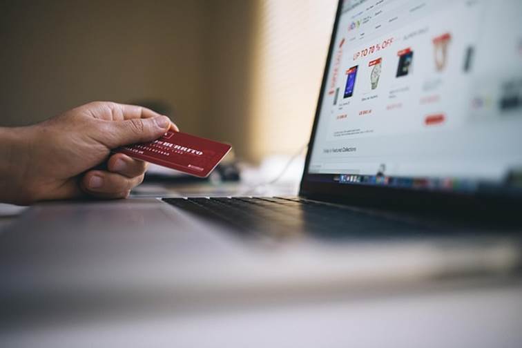Ruka držící červenou platební kartu před notebookem s otevřeným e-shopem se slevami, ilustrace online nákupu.